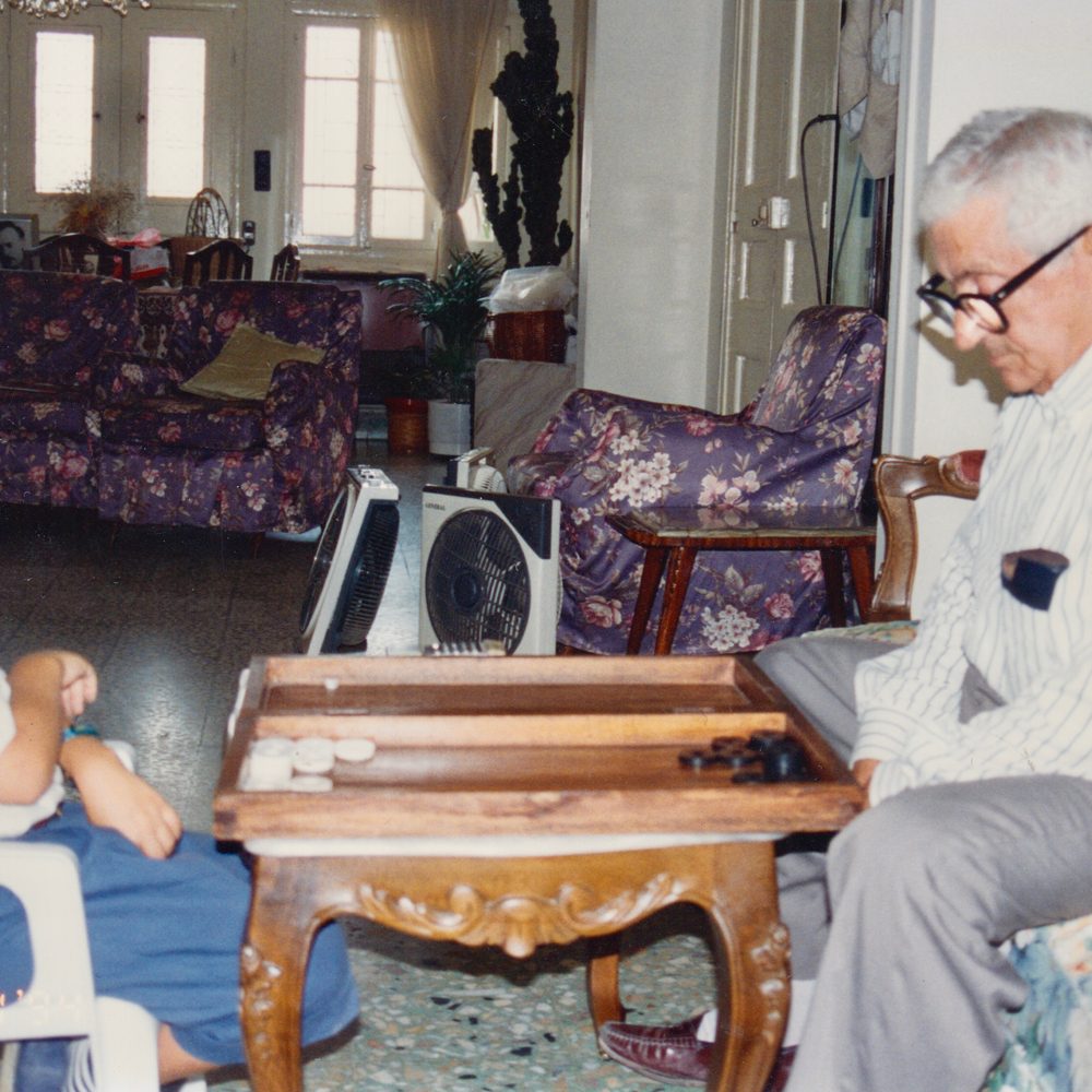 La photo est celle d'un amateur : un peu floue, mal cadrée. Au premier plan, une table de shesh besh en bois, sur laquelle sont posés des pions. À droite, un vieil homme à lunettes, aux cheveux blancs, regarde ses pions noirs. Il porte une chemise blanche à rayures, un pantalon de costume gris et des mocassins. À gauche, assis devant les pions blancs, on aperçoit les jambes et les bras d'un enfant. À l'arrière-plan, une salle de séjour : canapé et fauteuil à motifs fleuris, climatiseurs, table et chaises en bois verni, fenêtres aux voilages blancs.