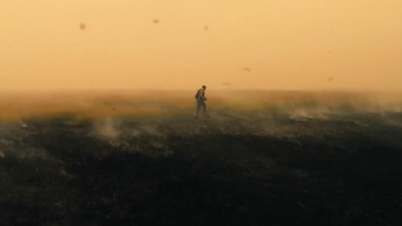 Une petite silhouette avance au milieu d'une steppe, remplie de cendres et de fumée, devant un ciel orange.