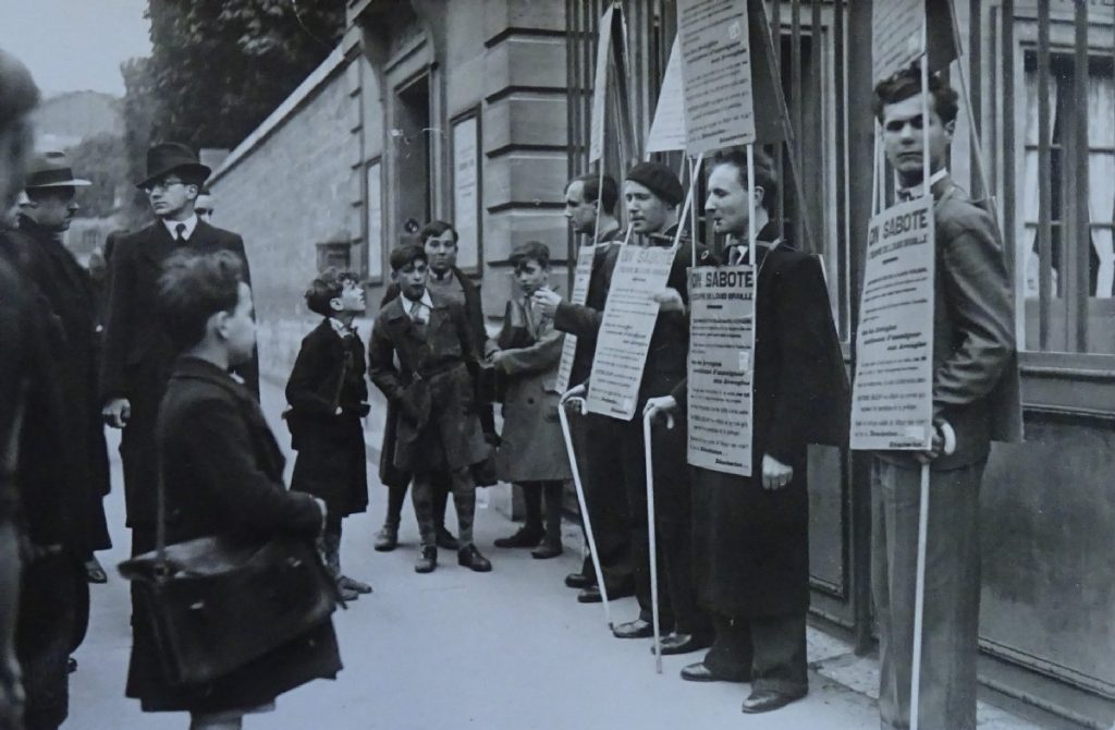 Photographie en noir et blanc : une dizaine d'hommes et jeunes garçons, debout à gauche et au fond, observent, debout à droite, quatre jeunes hommes aveugles manifestant devant une grille. Ils portent chacun une canne blanche et une triple pancarte formant un triangle au-dessus de leur tête, sur leur torse et sur leur dos, sur laquelle on distingue les mots « On sabote l'œuvre de Louis Braille ».