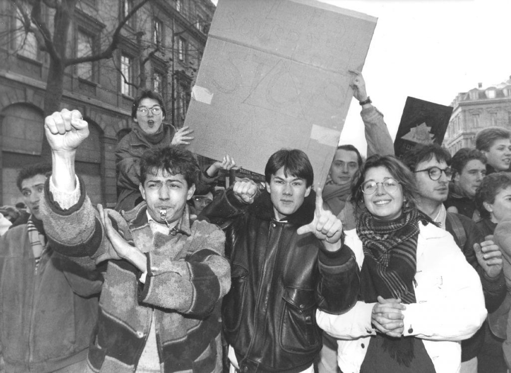 Photographie en noir et blanc d'une dizaine de jeunes hommes et femmes manifestant. Au premier plan, deux d'entre eux font face à l'appareil et signent le logo de l'association Sourds en colère, un triangle traversé par un poing. Derrière, deux autres tiennent une pancarte en carton sur laquelle on distingue les mots « Les sourds en colère. Stop !! Non à l'implant ».