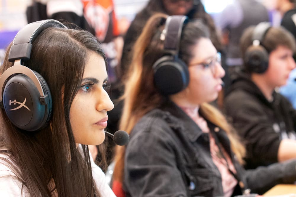 Une jeune femme brune aux cheveux longs, portant un casque audio avec un micro intégré, regarde droit devant elle d'un air déterminé. Dans son iris, se reflète un écran. En arrière-plan, flou, une autre jeune femme et un jeune homme sont dans la même position.