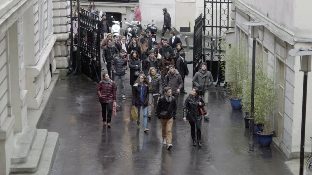 Une petite foule de jeunes gens pénètre dans une cour en passant une grande grille en fer forgée ouverte.