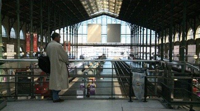 Surplombant les quais d'une gare, une personne de dos, en imperméable, regarde les voies accoudée à une rambarde.