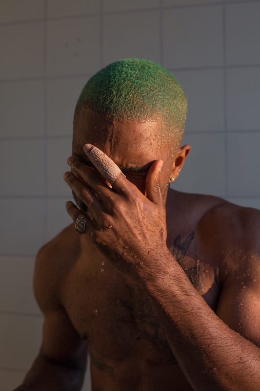 Frank, in the shower, de Wolfgang Tillmans, réalisé en 2015, est un portrait puissant capturant un homme torse nu sous une douche carrelée de blanc. L’homme, aux cheveux teints en vert vif, est saisi dans un moment d'intimité : il se couvre partiellement le visage de la main, les gouttes d’eau perlant sur sa peau sombre et musclée. Un bandage clair entoure l’un de ses doigts, ajoutant une note de fragilité à la force de sa posture. La lumière naturelle caresse son corps, soulignant la texture humide de sa peau et révélant subtilement un tatouage sur son torse. L’expression du sujet, bien que partiellement dissimulée, suggère une émotion profonde, entre pudeur, recueillement et intensité intérieure. Par cet instantané, Tillmans explore la vulnérabilité masculine et la beauté brute, dans un dialogue nuancé entre présence physique et intériorité émotionnelle.