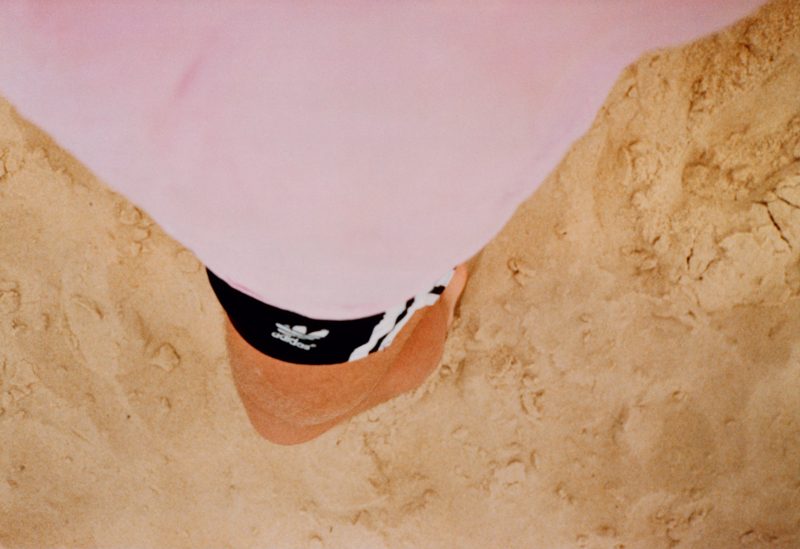 Lacanau (self) de Wolfgang Tillmans, réalisée en 1986, propose une vue subjective et intimiste ; un moment de détente à la plage. Photographiée depuis un angle plongeant vers le sol, l’image montre les jambes nues de l’artiste partiellement couvertes par un short noir Adidas, surmonté d'un t-shirt rose pastel qui envahit la partie supérieure du cadre. Le sable clair, parsemé d’empreintes et de textures naturelles, occupe tout l’arrière-plan. L'absence de repères directs (horizon, mer) recentre l’attention sur l’immédiateté de l'expérience corporelle et sensorielle. L'image évoque une sensation d’ancrage et de simplicité, traduisant l’esprit de liberté, de chaleur estivale et d’introspection douce qui caractérise souvent le travail de Tillmans. Cette photographie témoigne aussi d’une approche documentaire et autobiographique précoce, où l’instant personnel devient universel.