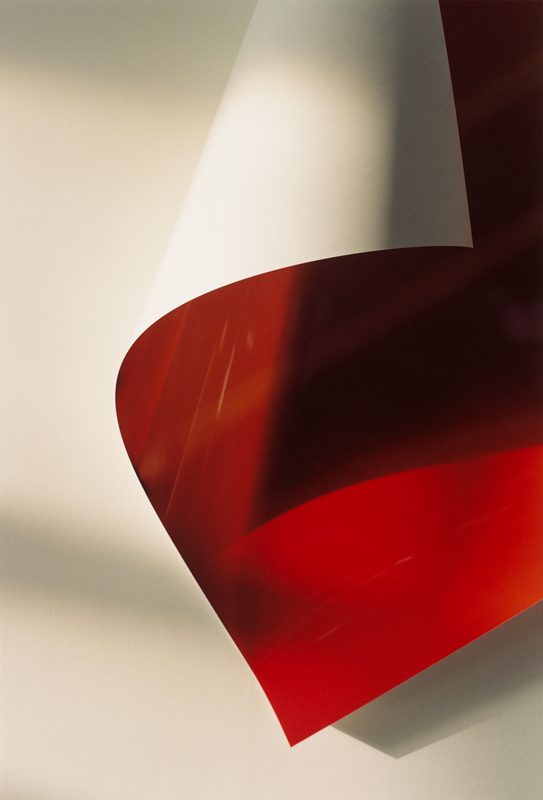 Paper drop 22, de Wolfgang Tillmans, réalisée en 2001, présente une composition épurée où une feuille de papier se déploie en une élégante courbe suspendue. La partie supérieure de la feuille reste blanche, tandis que sa face inférieure, d’un rouge éclatant et légèrement translucide, capte et reflète la lumière ambiante. Les jeux d’ombres portées sur le mur neutre renforcent la tridimensionnalité du papier, accentuant l’illusion d’un mouvement délicat figé dans l’instant. Les lignes douces et les transitions subtiles entre les tons créent une esthétique à la fois minimaliste et sensuelle. Tillmans explore ici les propriétés physiques du papier — sa légèreté, sa flexibilité, sa capacité à modeler l'espace — tout en transcendant la nature ordinaire de l’objet pour en révéler une beauté sculpturale. La maîtrise de la lumière naturelle joue un rôle clé, conférant à la scène une douceur presque tactile.