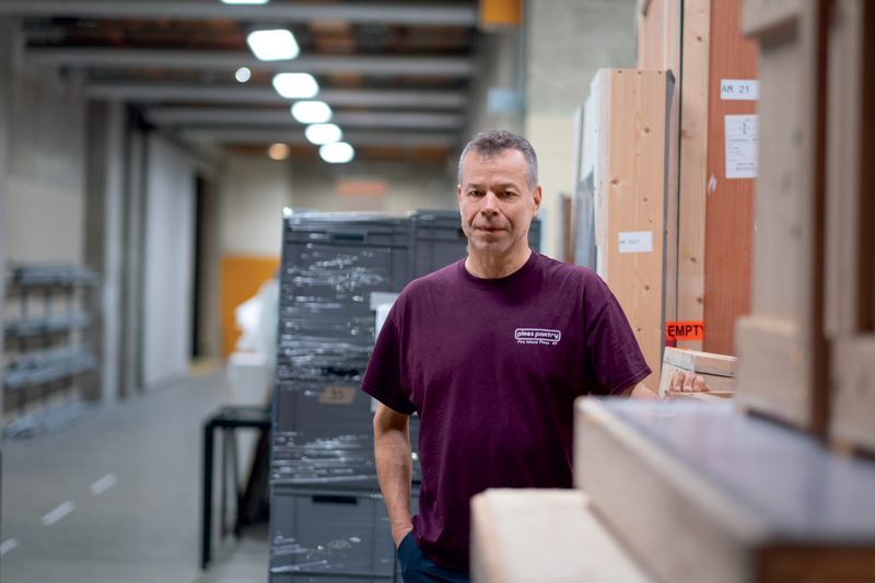 Wolfgang Tillmans est photographié dans un espace logistique du Centre Pompidou, au milieu des caisses de transport destinées aux œuvres d'art. Vêtu d’un t-shirt bordeaux marqué « Démé à Paris », il adopte une posture naturelle, une main dans la poche, l’autre posée légèrement sur une caisse en bois. Son regard, serein et attentif, traduit une implication directe dans les opérations de montage de son exposition. L’environnement industriel, avec ses caisses en bois étiquetées « Fragile », ses rayonnages métalliques et ses lignes de fuite marquées par les plafonniers alignés, évoque l'envers discret mais fondamental du monde muséal. Les tons neutres de béton brut, bois clair et emballages plastiques participent à instaurer une atmosphère méthodique, révélant la dimension matérielle de la création et de l'exposition artistique.