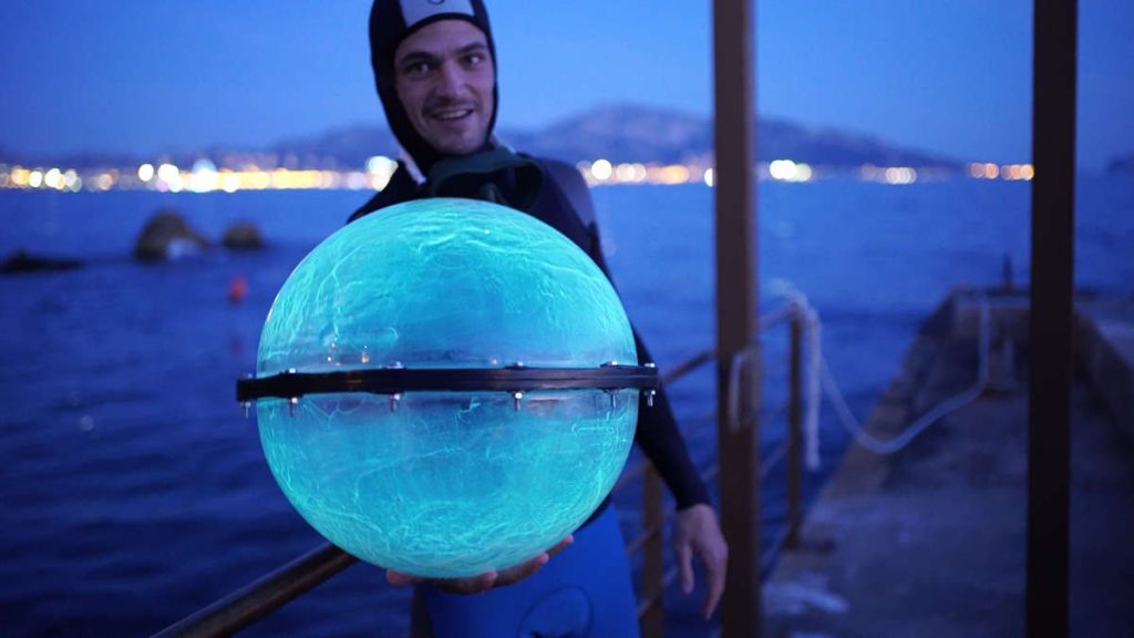 Jeremy Bruguidou en tenue de plongée, une de ses sphères bioluminescentes à la main. La sphère en gros plan fait environ 60 cm de diamètre