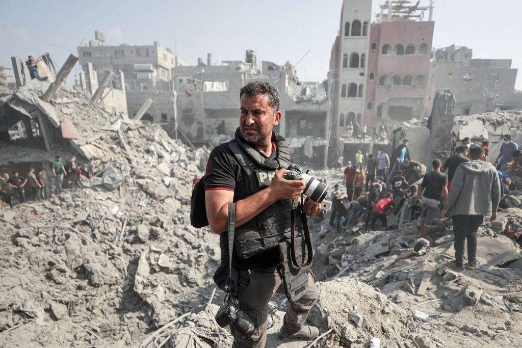 Le journaliste porte un gilet par-balles Press. Il se tient sur un monticule de gravats, appreil photo en main. En arrière plan, une ville en ruine et sur le côté des personnes avancent en file indienne, chargées de bagages.