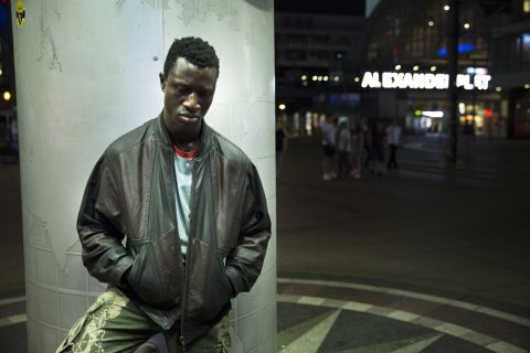 Image tirée du film Berlin Alexanderplatz de Burhan Qurbani : un homme adossé à une colonne, les mains dans les poches de sa veste, avec en arrière-plan un espace urbain de nuit avec le mot Alexanderplatz en néons blancs