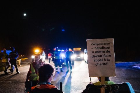 Photographie en couleur prise de nuit : au premier plan, deux personnes en fauteuil roulant, au milieu d'une route, vues de dos, avec une pancarte « Compensation de handicap = marre de devoir faire appel à la charité ! ». À l'arrière-plan, des véhicules aux phares allumés et des personnes debout portant des gilets jaunes ou des uniformes de gendarmerie.