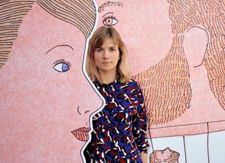 Photo, portrait couleur de Manon Fayolle. On ne voit que son visage et son buste. C’est une jeune femme blonde, aux yeux clairs et aux cheveux mi-longs. Elle se tient debout dans un décor composé de ses dessins représentant deux visages.